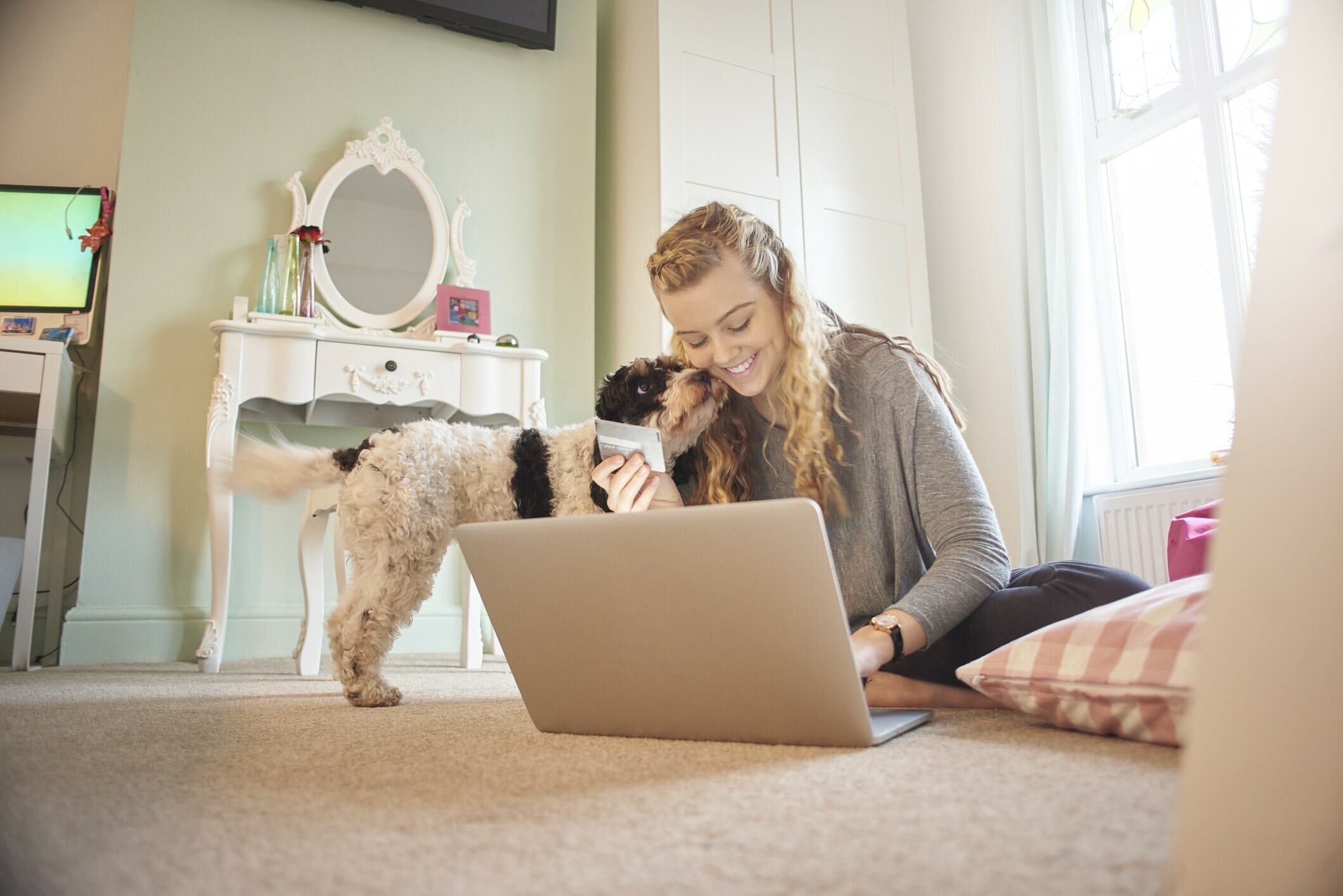 curly haired happy woman sits on bedroom floor with her dog laptop, learning about credit